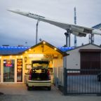 Erik Tham - Retired Supersonic Birds: Barely visible behind a pizza restaurant and industrial buildings, a discarded Concorde supersonic passenger aircraft rests in peace at the rooftop of the museum for technology in Sinsheim, Germany